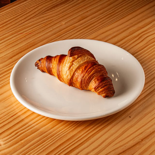 Croissant dorado y crujiente servido en un plato blanco sobre una mesa de madera.