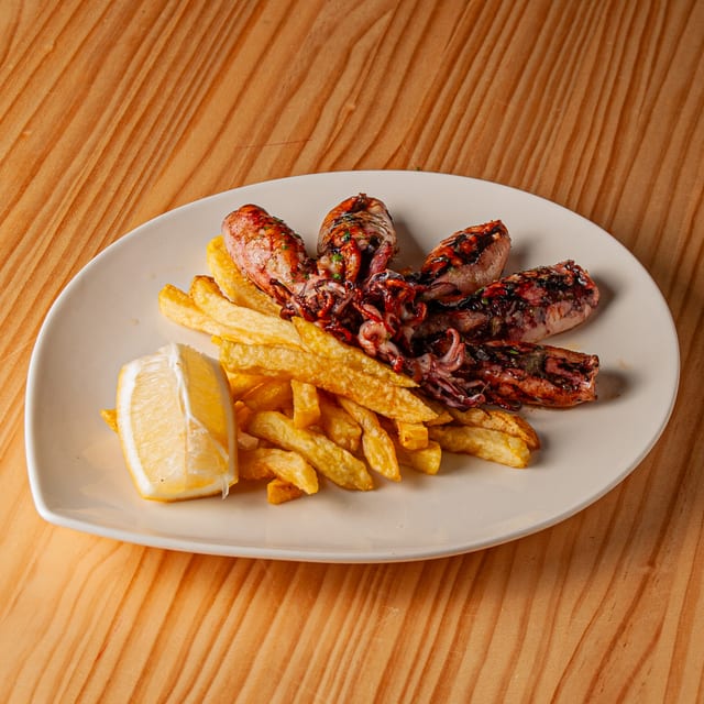 Plato de chipirones a la plancha acompañados de patatas fritas y un gajo de limón sobre mesa de madera.