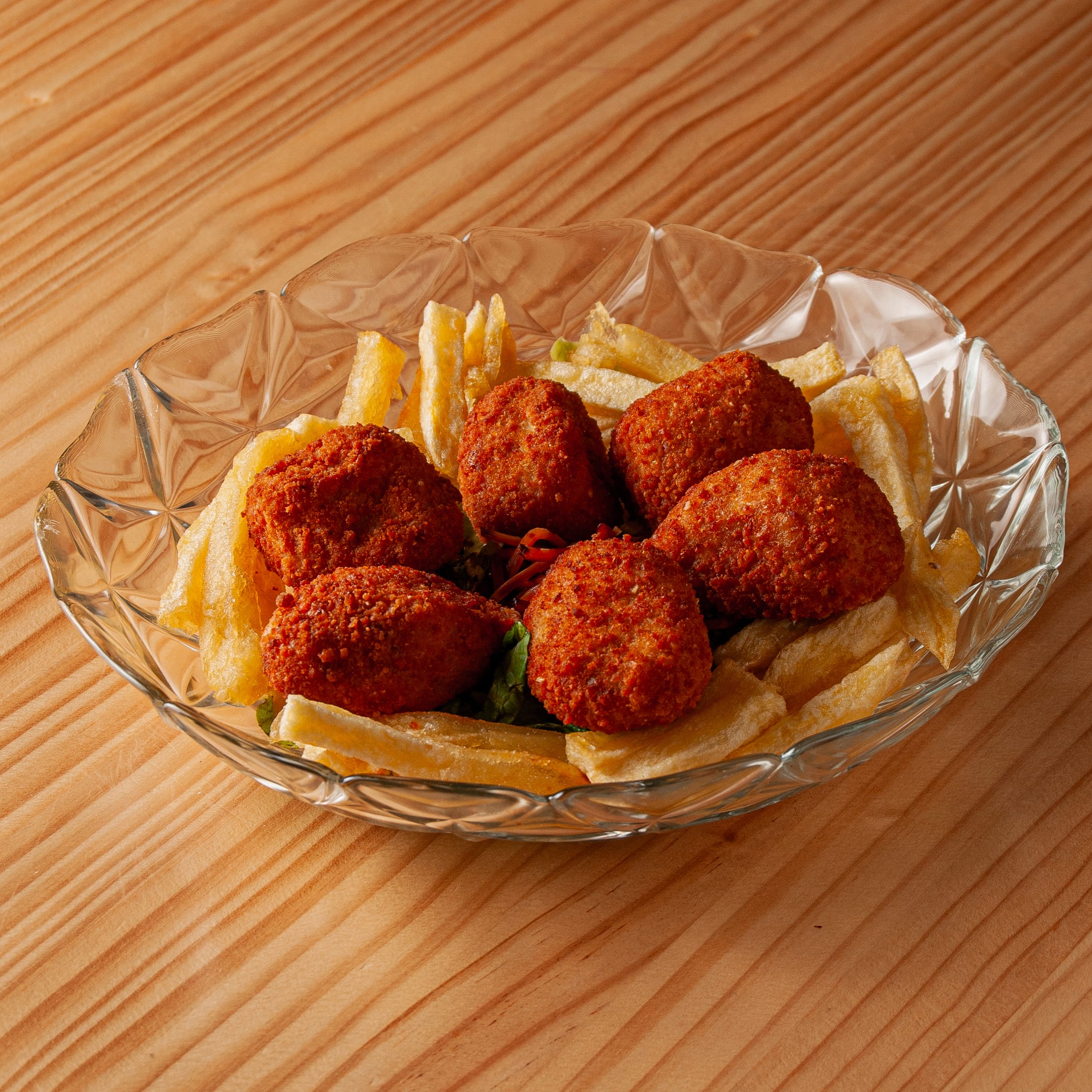 Plato de cristal con varias croquetas de jamón doradas y crujientes acompañadas de patatas fritas sobre una mesa de madera.