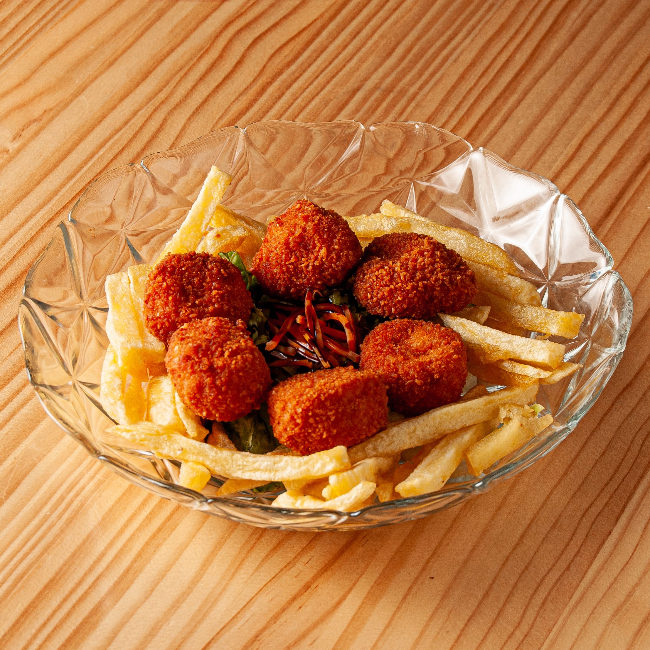 Plato de croquetas de merluza doradas y crujientes servidas sobre una cama de patatas fritas en un bol de cristal sobre una mesa de madera.
