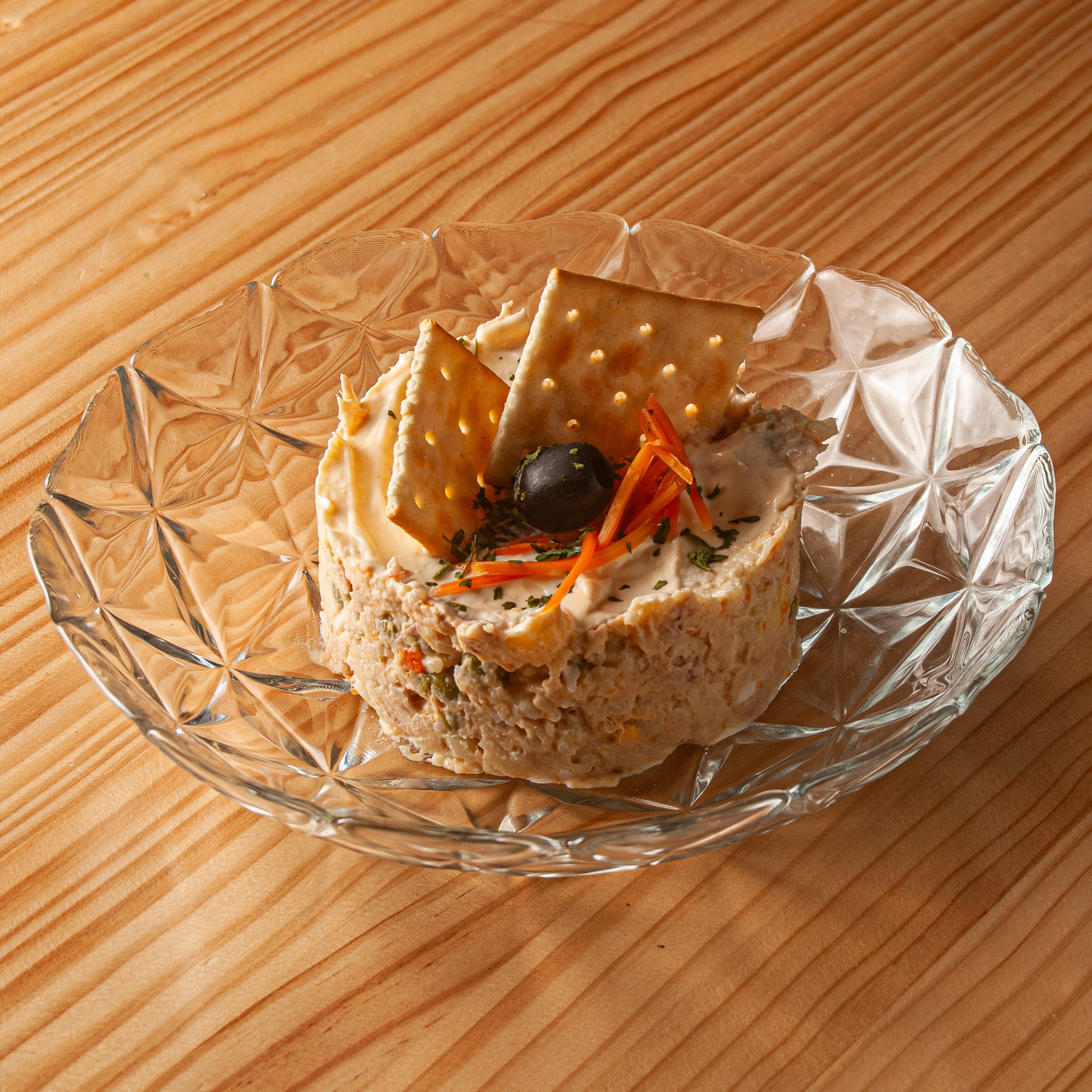 Porción de ensaladilla rusa servida sobre un plato de cristal con diseño tallado, decorada con una aceituna negra, tiras de zanahoria y dos galletas saladas, sobre una mesa de madera.