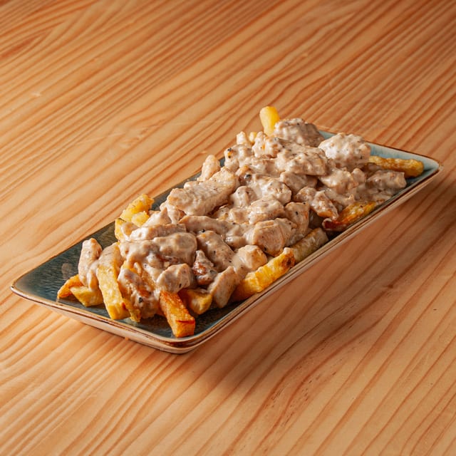 Plato rectangular con patatas fritas cubiertas de trozos de pollo salteados con abundante salsa cremosa de queso, sobre una mesa de madera.