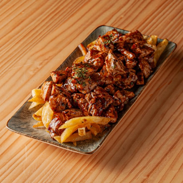 Plato rectangular con raxo de cerdo troceado bañado en salsa y servido sobre una cama de patatas fritas sobre mesa de madera.