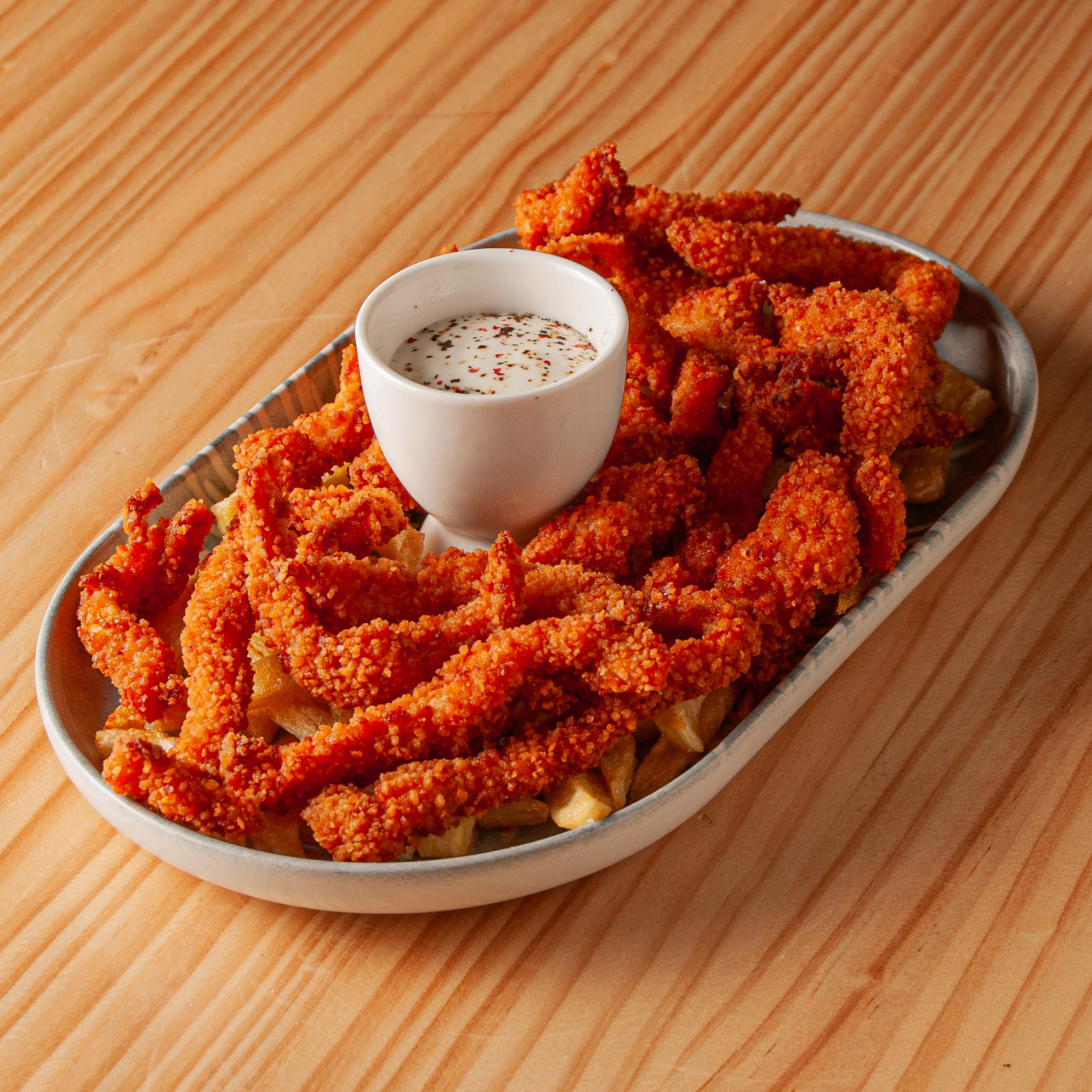 Tiras de pollo crujientes y doradas servidas sobre una cama de papas fritas en un plato ovalado con una pequeña salsera de salsa cremosa con especias sobre una mesa de madera.