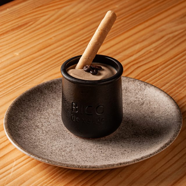 Postre de café servido en un pequeño recipiente de cerámica negra con la marca Bico de Xeado, decorado con granos de café y un barquillo crujiente sobre un plato de piedra moteado en una mesa de madera.