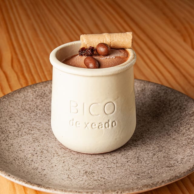 Helado de chocolate servido en un tarro de cerámica con la marca Bico de xeado, decorado con un barquillo y granos de café sobre un plato rústico.