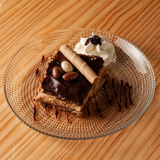 Porción de tarta de la abuela con cobertura de chocolate, decorada con un barquillo, pequeños huevos de chocolate y una nube de nata montada sobre un plato de cristal con textura.