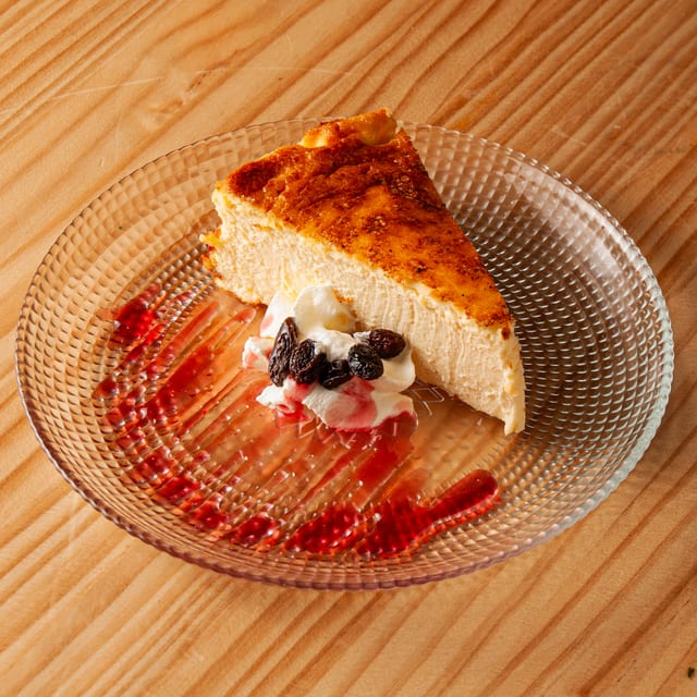 Porción de tarta de queso horneada con superficie dorada, servida con una nube de nata montada, pasas y un sirope de frutos rojos sobre un plato de cristal texturizado en una mesa de madera.