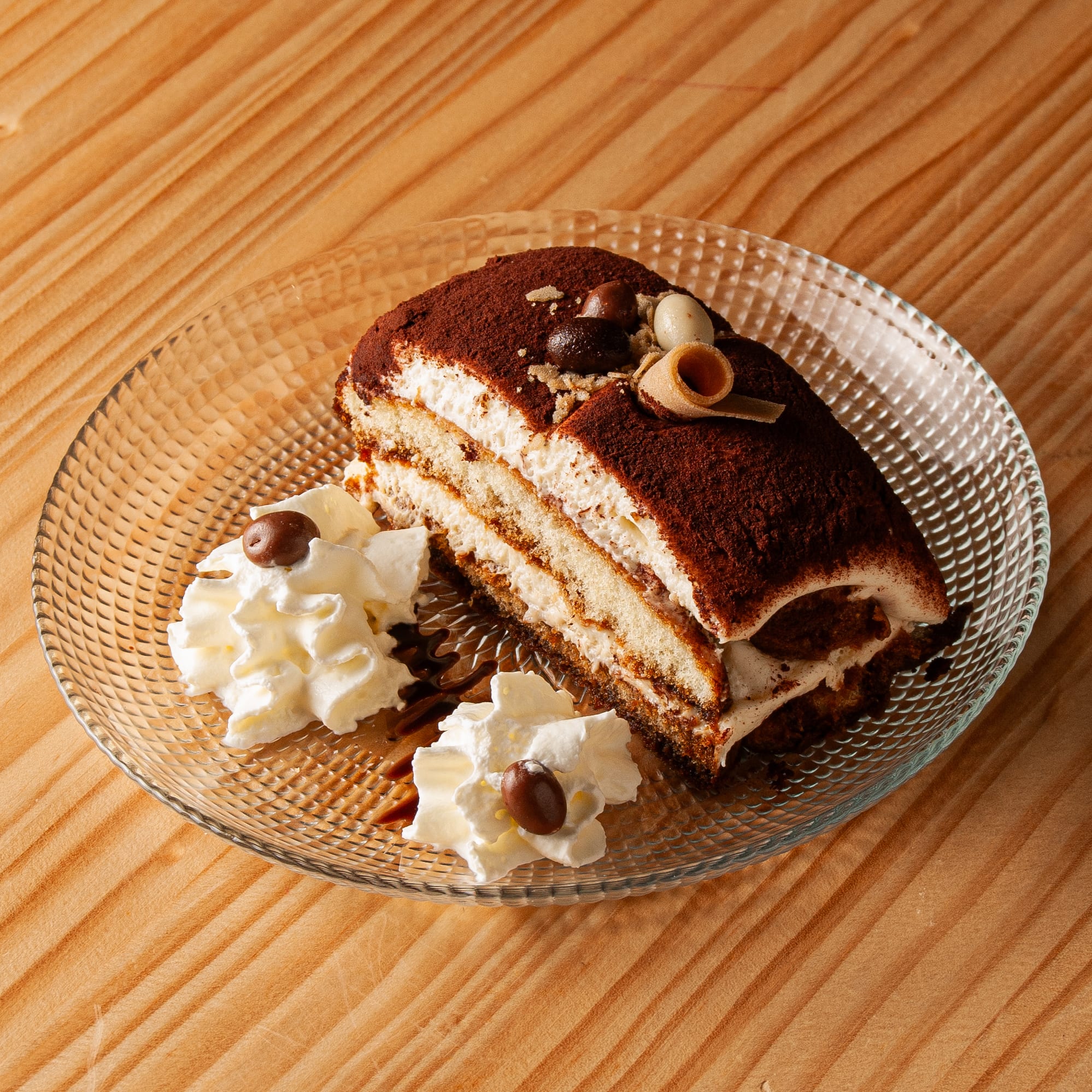 Porción de tiramisú espolvoreada con cacao, decorada con un barquillo y grageas de chocolate sobre un plato de cristal, acompañada de dos montículos de nata montada con granos de café de chocolate.