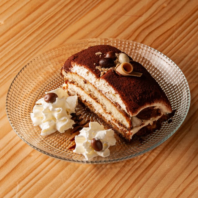 Porción de tiramisú espolvoreada con cacao, decorada con un barquillo y grageas de chocolate sobre un plato de cristal, acompañada de dos montículos de nata montada con granos de café de chocolate.