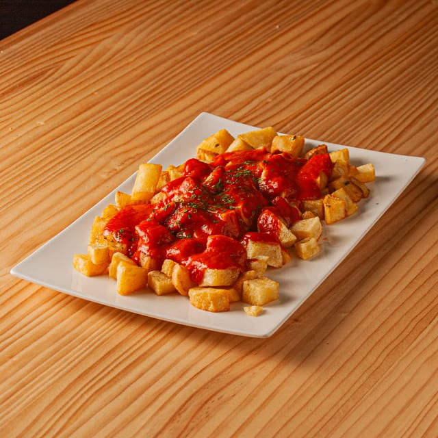 Ración de patatas bravas doradas con salsa roja picante y un toque de hierbas aromáticas sobre plato blanco rectangular, apoyado en una mesa de madera clara.