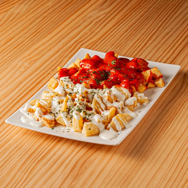 Ración de patatas bravioli sobre plato rectangular blanco, con una mitad cubierta de salsa brava roja y la otra mitad de alioli, espolvoreadas con perejil fresco sobre una mesa de madera.