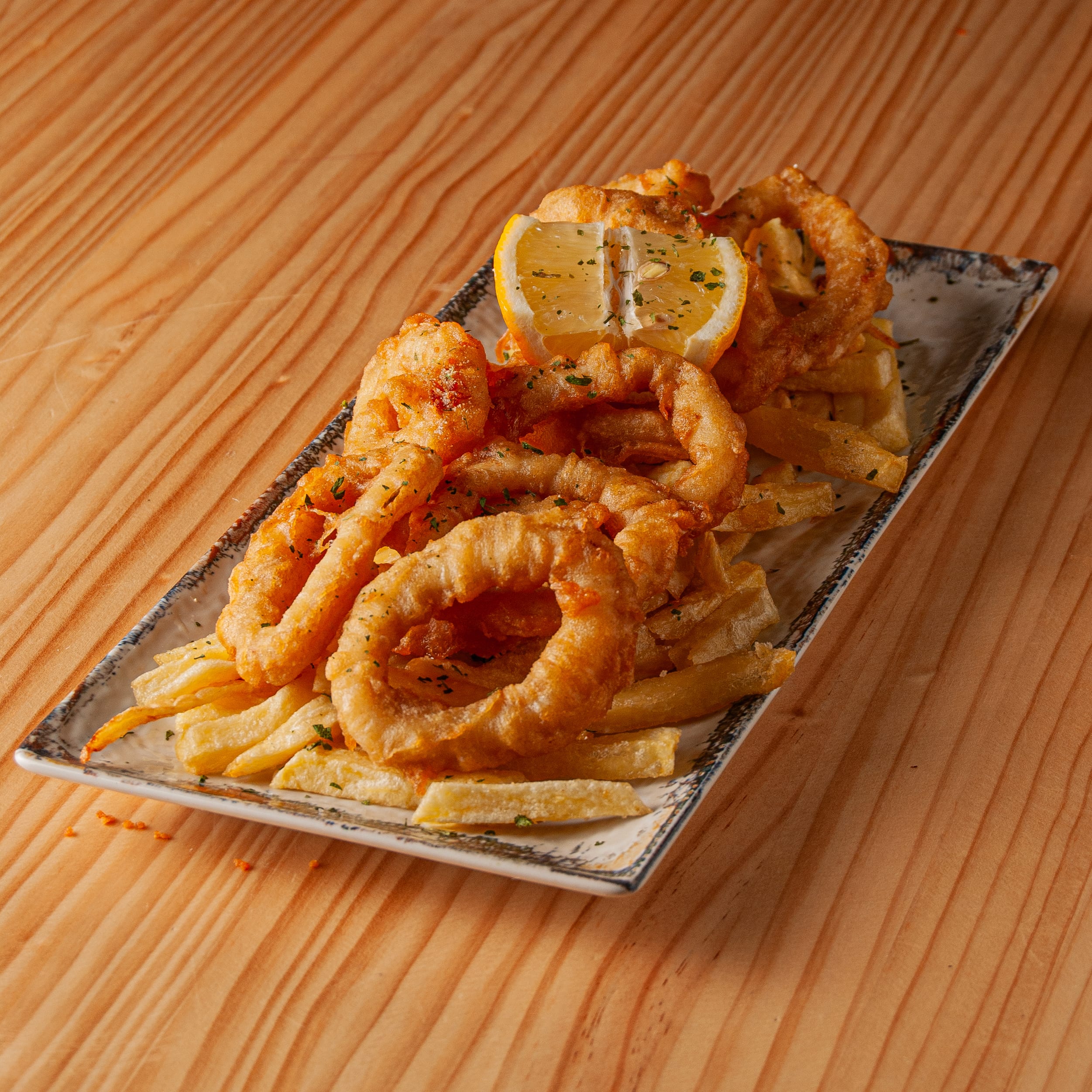 Ración de calamares a la romana crujientes servidos con patatas fritas y una rodaja de limón sobre una bandeja rectangular de estilo rústico sobre mesa de madera.