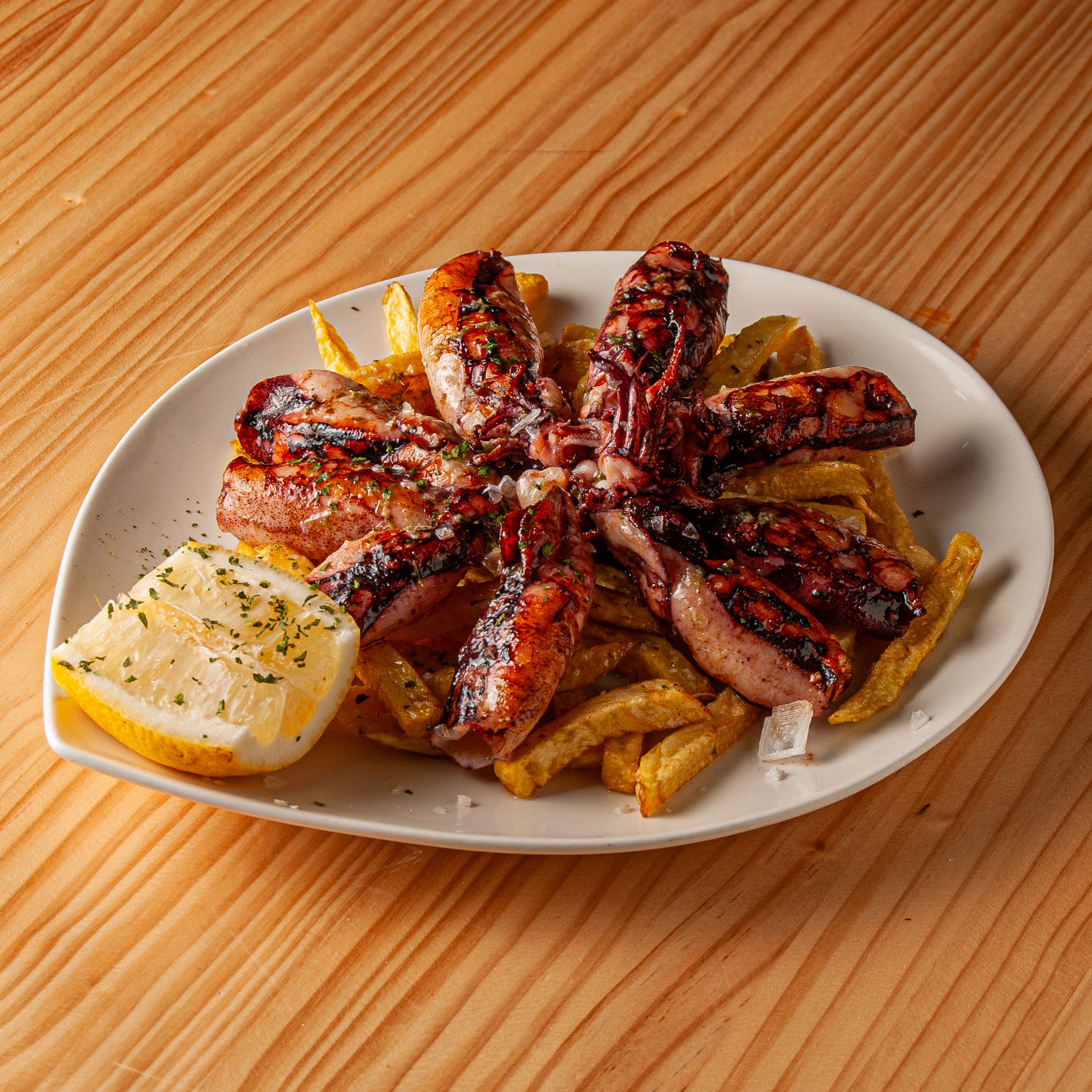 Ración de chipirones a la plancha sobre una cama de patatas fritas servida en un plato blanco con un gajo de limón y espolvoreada con perejil.