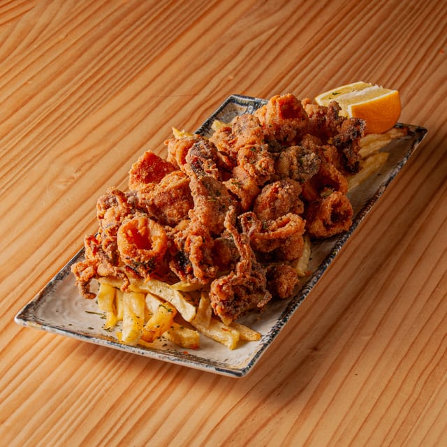 Ración de chipirones rebozados y fritos servidos sobre una cama de patatas fritas en una bandeja rectangular, acompañados con un gajo de limón sobre una mesa de madera.