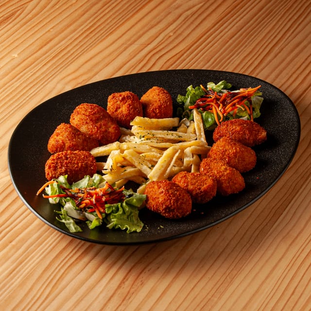 Ración de croquetas de jamón crujientes acompañadas de patatas fritas y una pequeña ensalada de lechuga y zanahoria sobre un plato negro ovalado en una mesa de madera.
