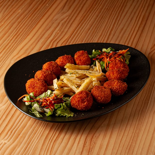 Ración de croquetas doradas de merluza acompañadas de patatas fritas y una guarnición de lechuga fresca con zanahoria rallada sobre un plato negro ovalado.
