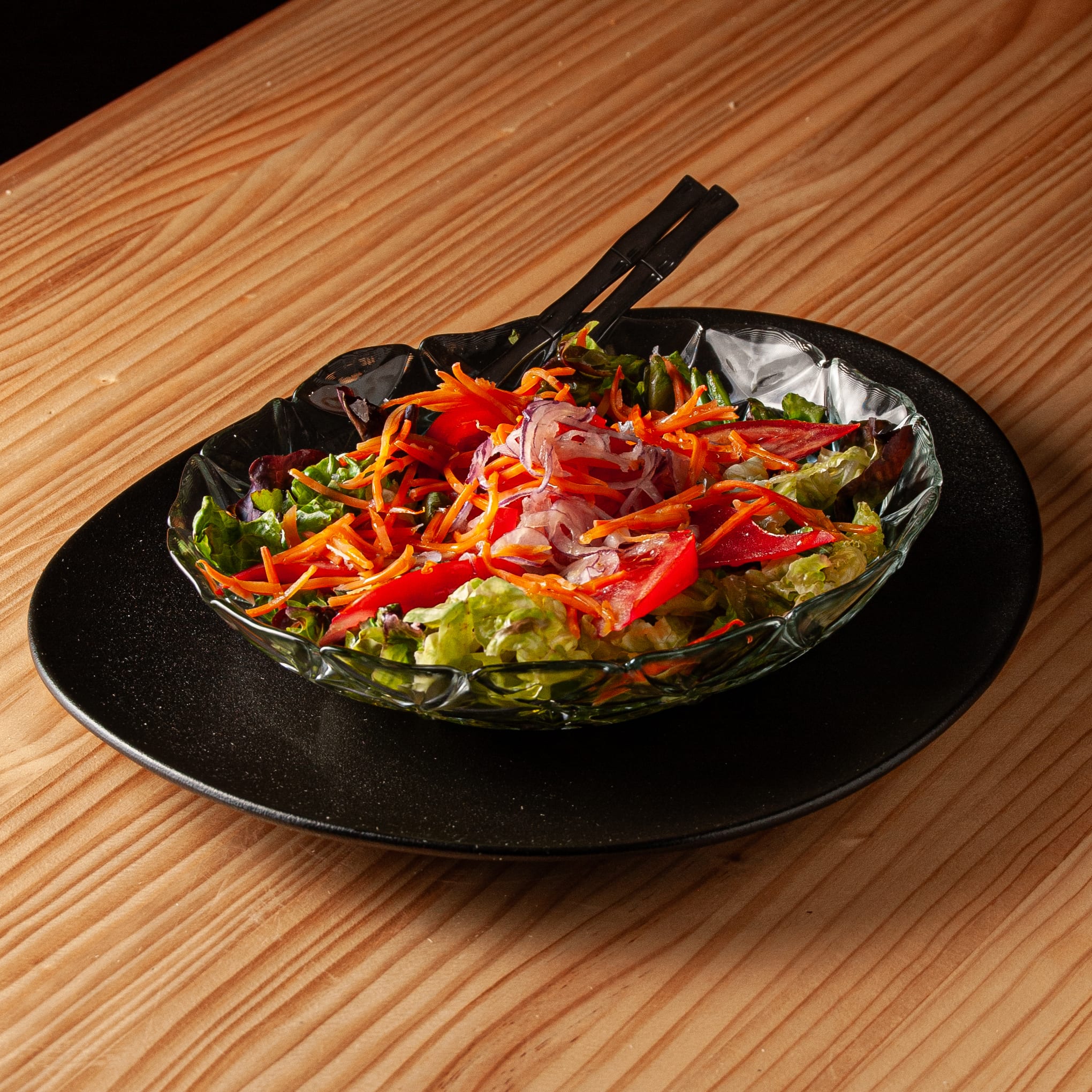 Ensalada clásica servida en un bol de cristal sobre un plato negro, compuesta por lechuga, zanahoria rallada, tomate y cebolla, con cubiertos negros descansando sobre la mezcla.