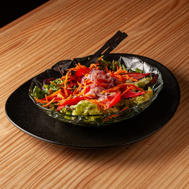 Ensalada clásica servida en un bol de cristal sobre un plato negro, compuesta por lechuga, zanahoria rallada, tomate y cebolla, con cubiertos negros descansando sobre la mezcla.