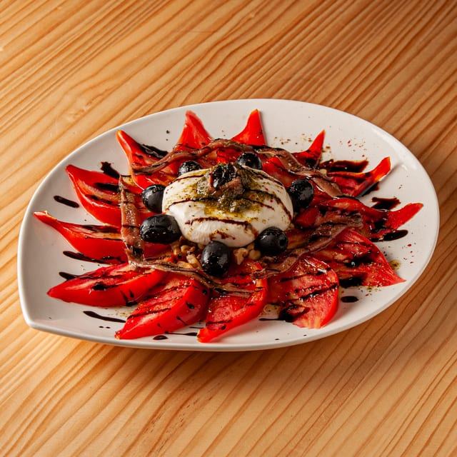 Ensalada rovelesca con tomate en rodajas, burrata central, aceitunas negras, anchoas y reducción de vinagre balsámico sobre plato blanco.