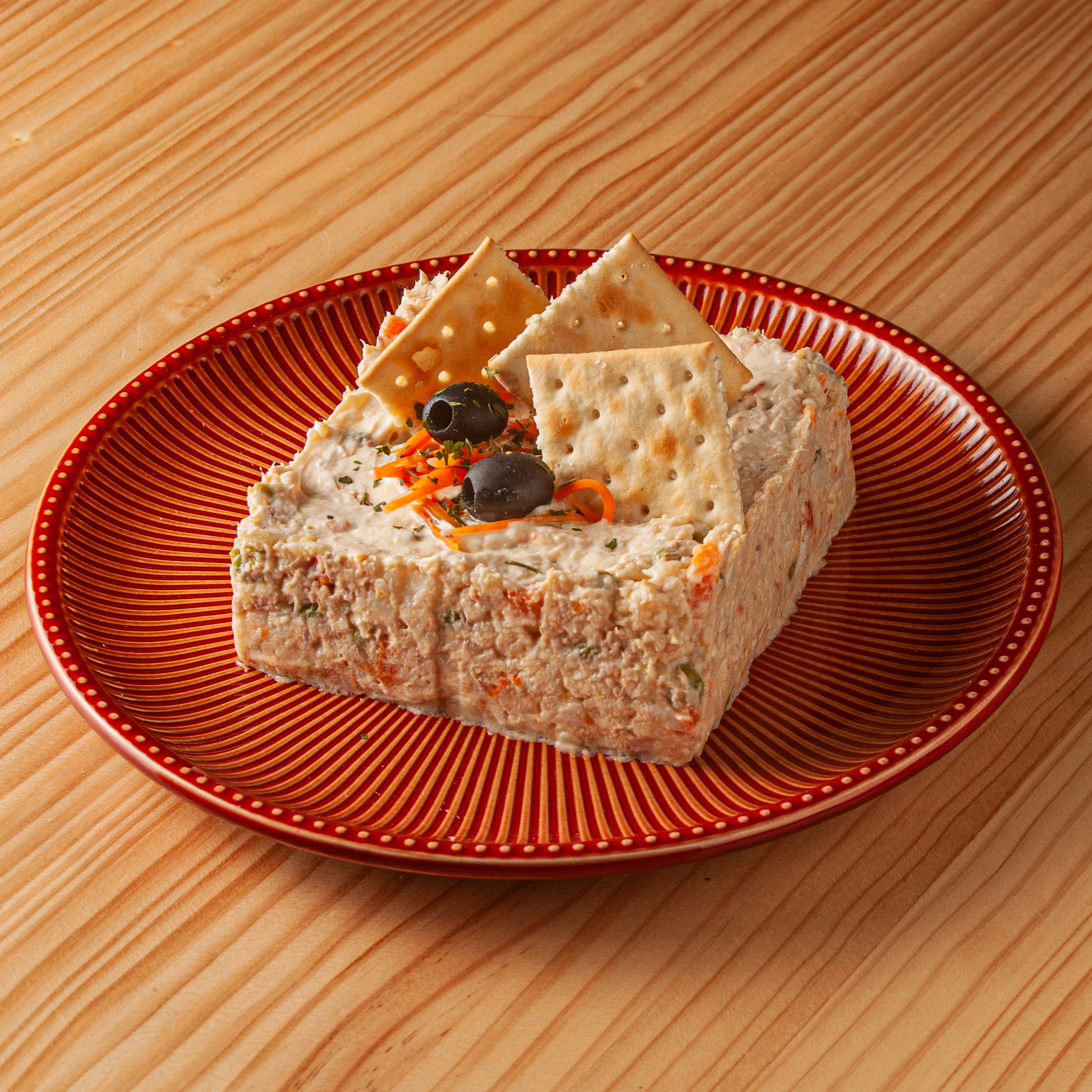 Ración rectangular de ensaladilla rusa servida en un plato rojo con relieve, decorada con aceitunas negras y galletas saladas sobre una mesa de madera.