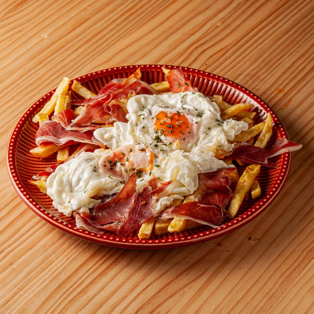 Plato de huevos fritos sobre una base de patatas fritas cortadas en bastones, acompañados con finas lonchas de jamón serrano y espolvoreados con hierbas aromáticas, servido en un plato rojo con relieve sobre una mesa de madera.