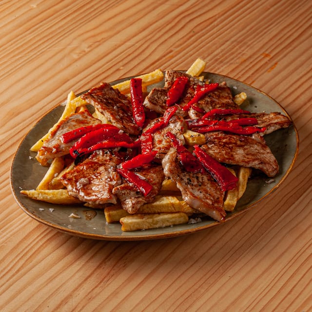 Ración de lomitos de cerdo a la plancha servidos sobre una cama de patatas fritas doradas y decorados con tiras de pimiento rojo asado en un plato de cerámica sobre mesa de madera.