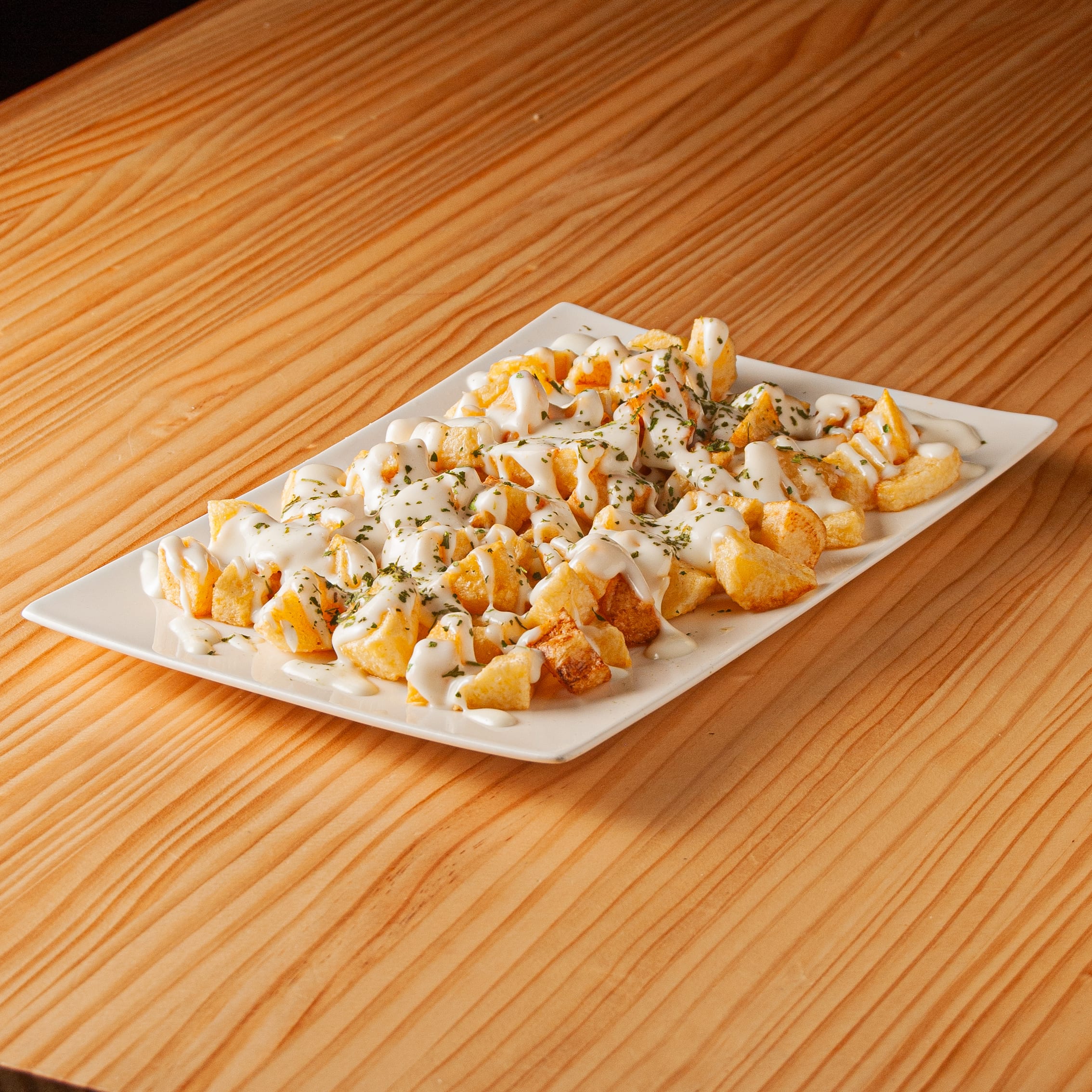 Ración de patatas fritas cortadas en dados con abundante salsa alioli y espolvoreadas con perejil fresco sobre un plato rectangular blanco.