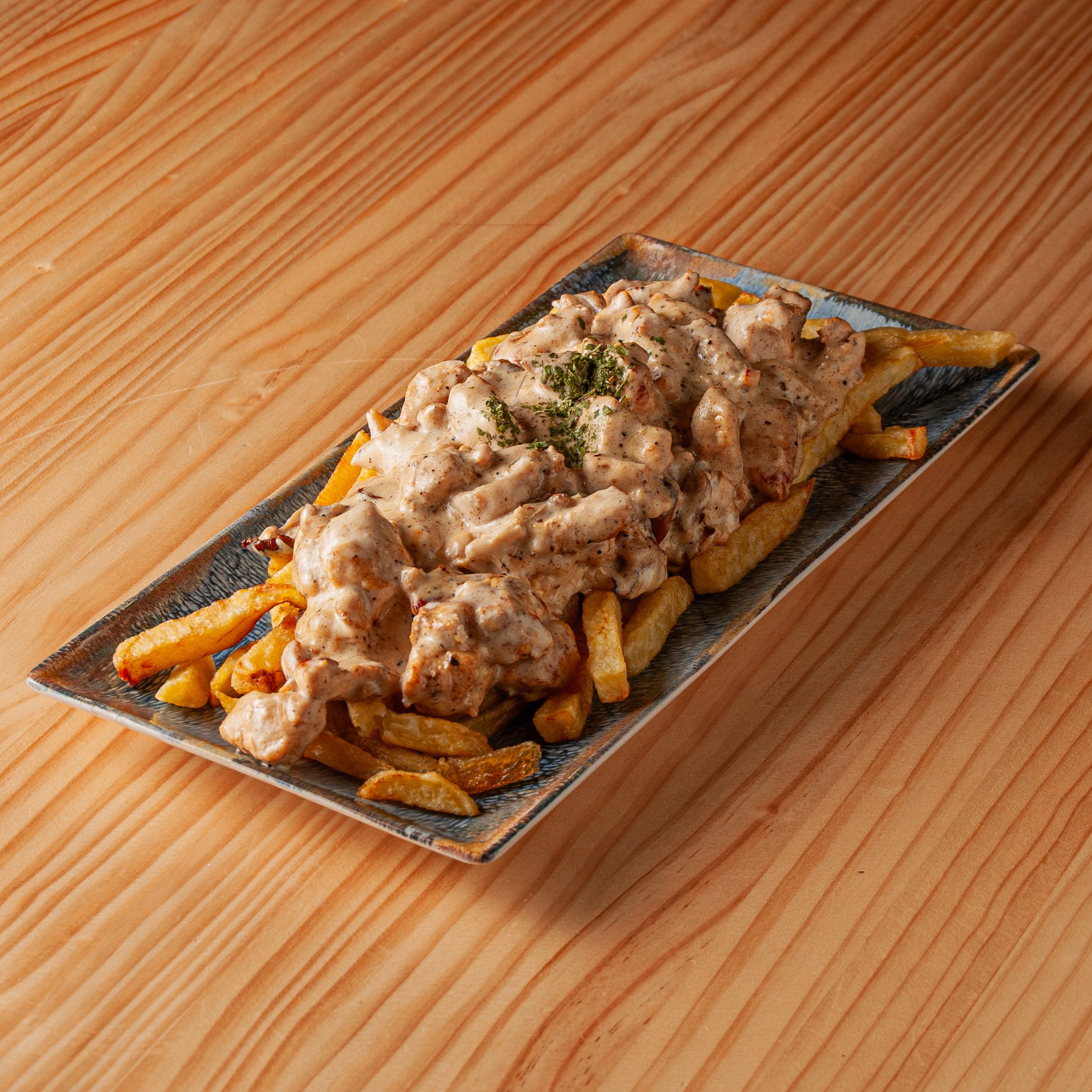 Ración de pollo con salsa cremosa de nata sobre una base de patatas fritas servido en un plato rectangular sobre una mesa de madera.
