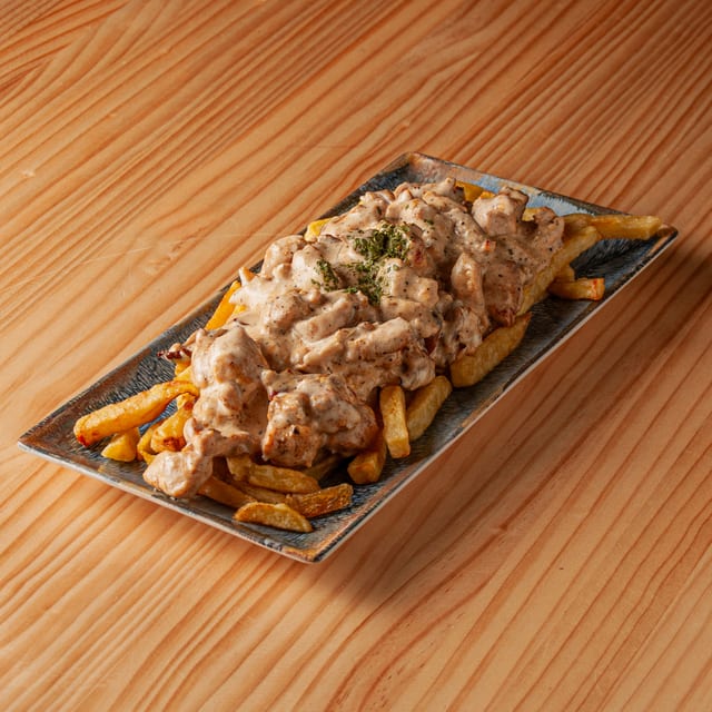 Ración de pollo con salsa cremosa de nata sobre una base de patatas fritas servido en un plato rectangular sobre una mesa de madera.
