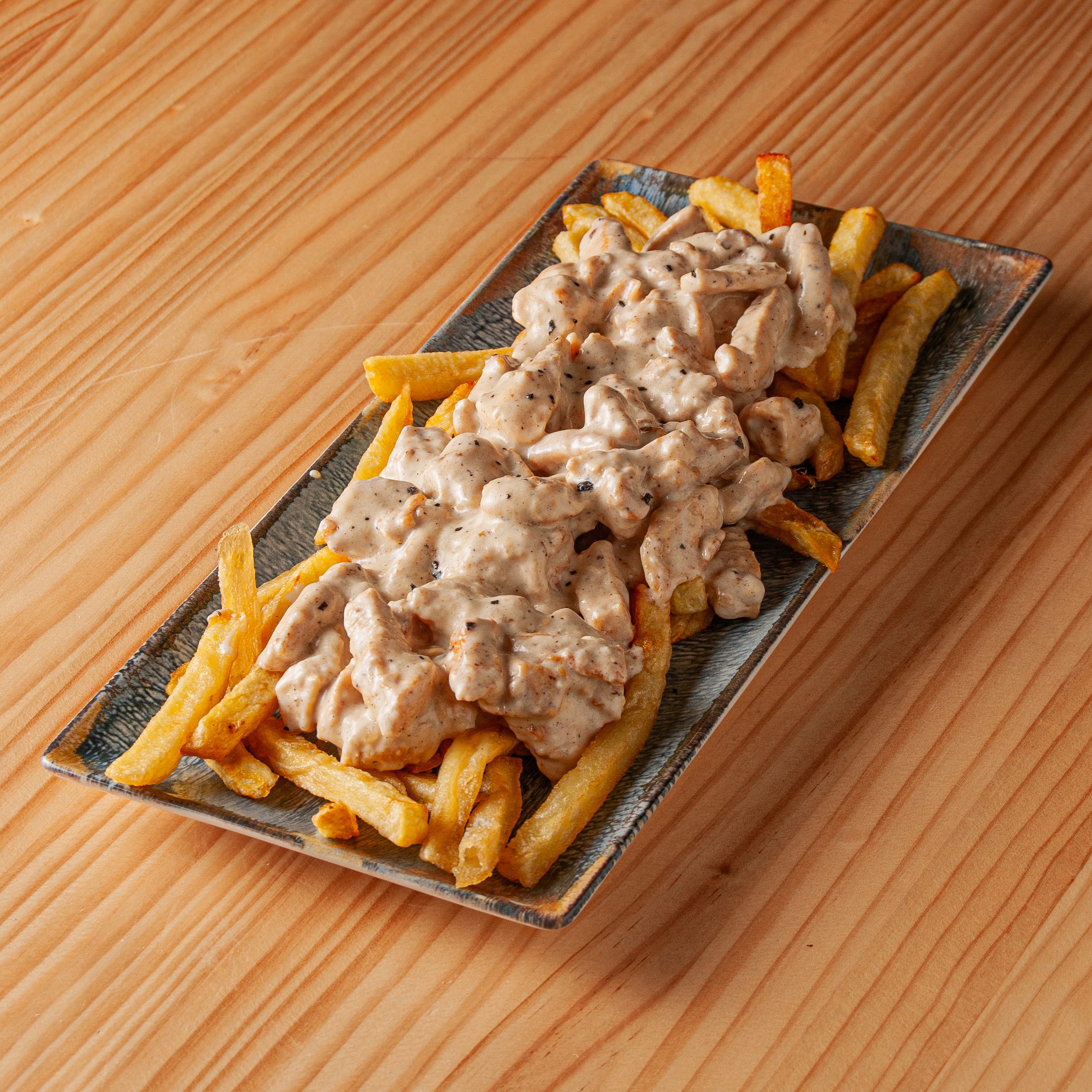 Ración de patatas fritas cubiertas con trozos de pollo en salsa cremosa de queso sobre bandeja rectangular de cerámica en mesa de madera.