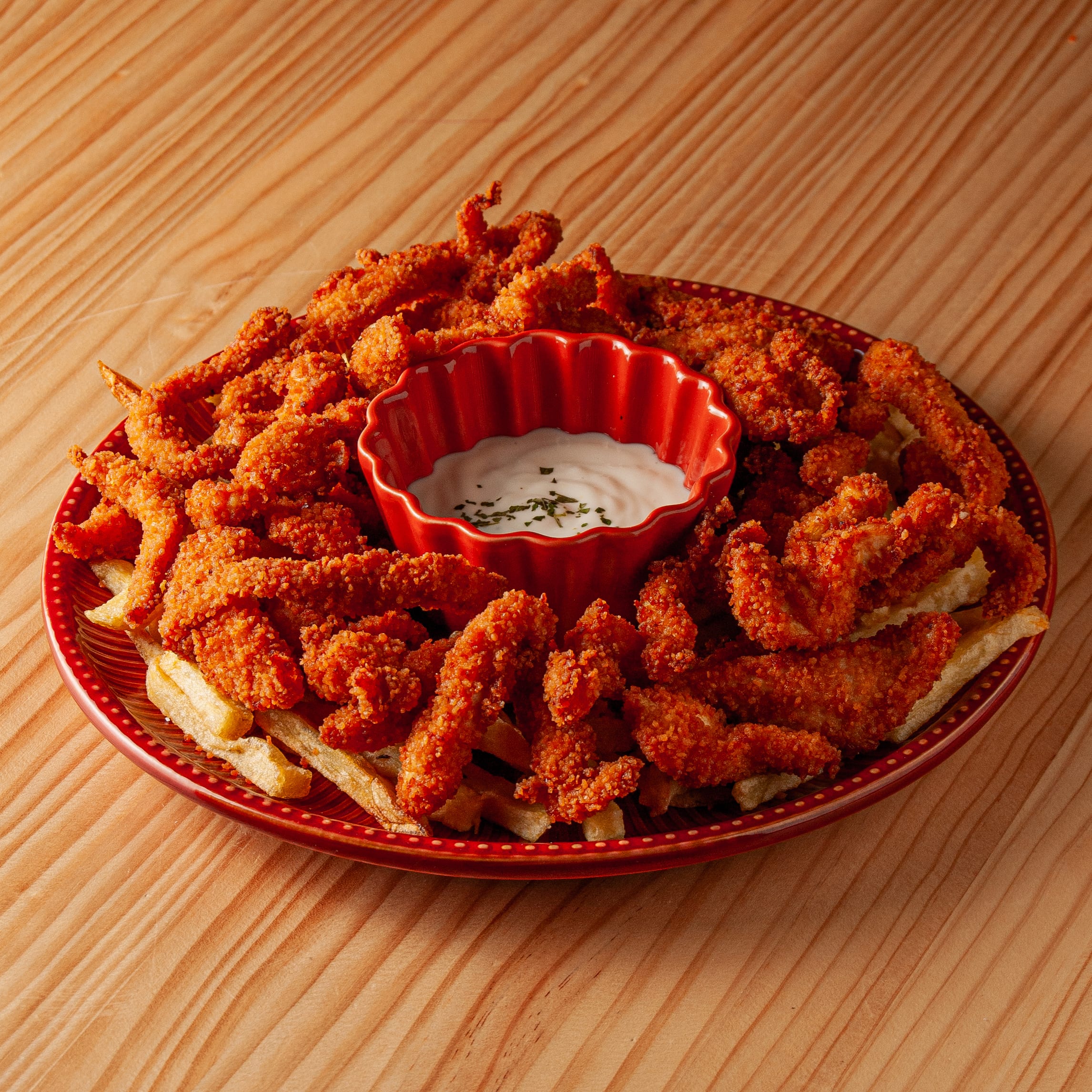 Plato con abundantes tiras de pollo crujientes y patatas fritas servido con un cuenco central de salsa cremosa sobre mesa de madera.