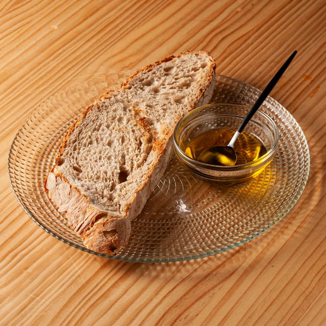 Rebanada de pan artesanal tostado servida en un plato de cristal con un cuenco de aceite de oliva virgen extra y una cuchara negra sobre una mesa de madera.