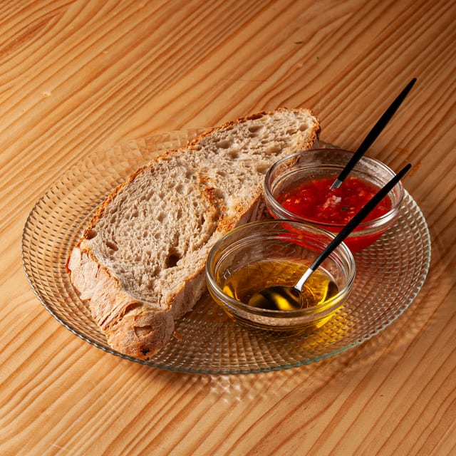 Rebanada de pan artesanal sobre plato de cristal acompañada de cuencos con tomate triturado y aceite de oliva virgen extra sobre mesa de madera.