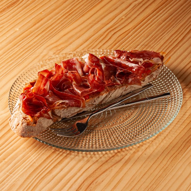 Tosta de pan artesano con jamón ibérico servida sobre un plato de cristal transparente con un tenedor, apoyada en una mesa de madera.