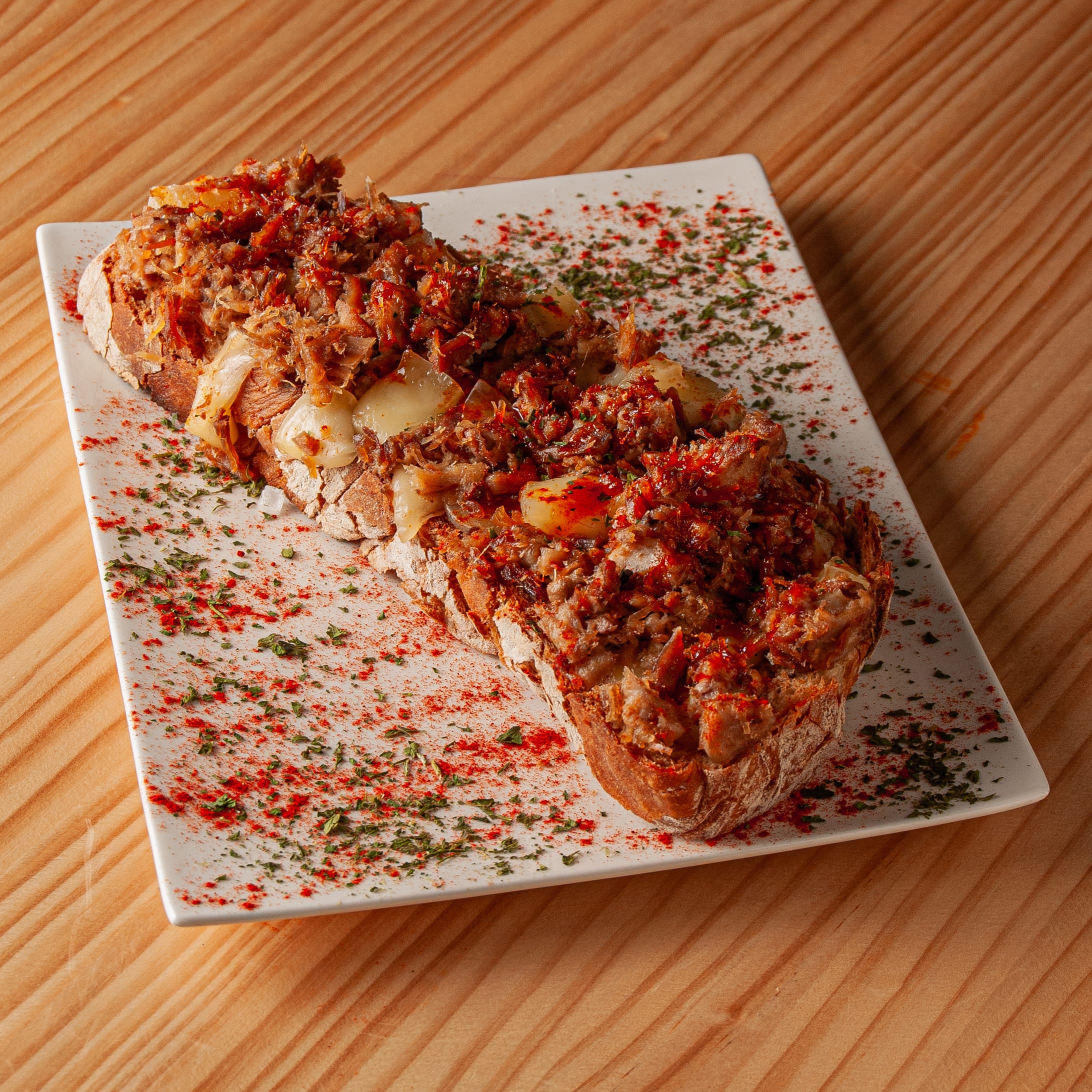 Tosta gallega con carne desmechada, queso fundido y pimentón sobre plato rectangular blanco en mesa de madera.