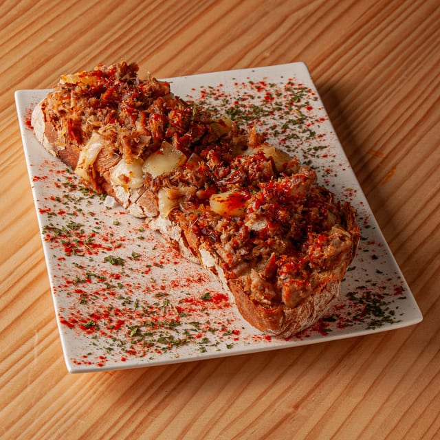 Tosta gallega con carne desmechada, queso fundido y pimentón sobre plato rectangular blanco en mesa de madera.