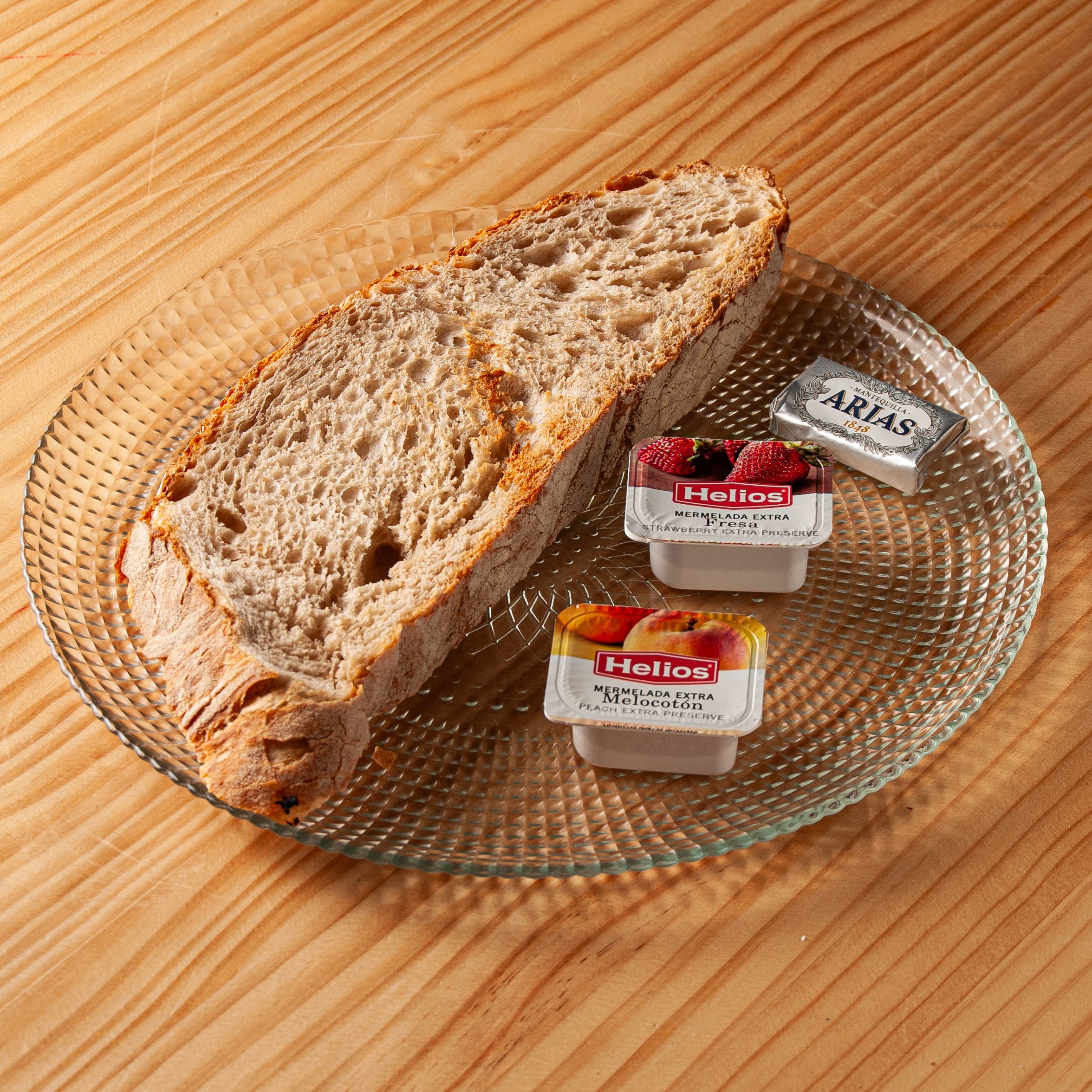 Rebanada de pan de masa madre en un plato de cristal con porciones individuales de mantequilla Arias y mermeladas Helios de fresa y melocotón sobre una mesa de madera.