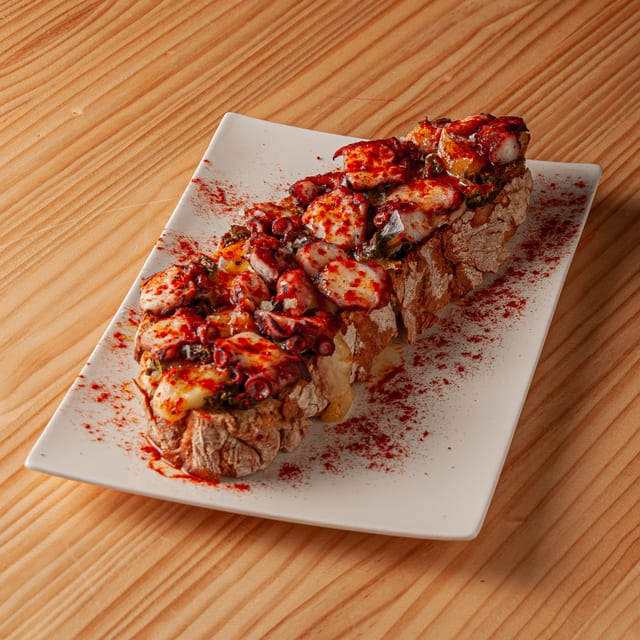 Tosta de pan artesano con pulpo a la gallega, aceite de oliva y pimentón espolvoreado, servida en plato rectangular blanco sobre mesa de madera.